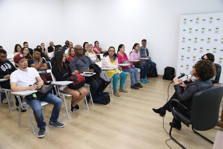 Foto do auditório da Fundação Dorina preenchido por cerca de 50 pessoas sentadas em cadeiras universitárias. À frente delas, também sentada, a escritora Cristiane Correa fala ao microfone.